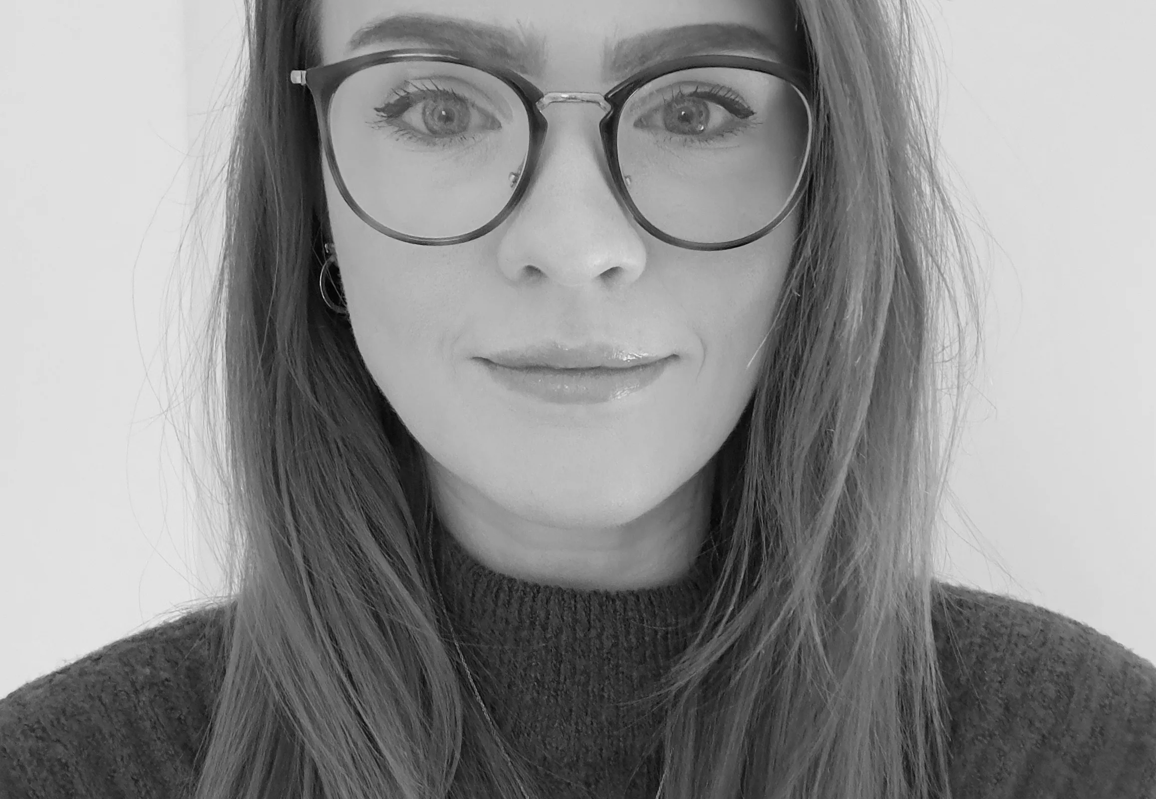 Black and white close-up of a person with long hair and glasses, wearing a dark sweater. The expression is neutral, creating a calm, focused tone.