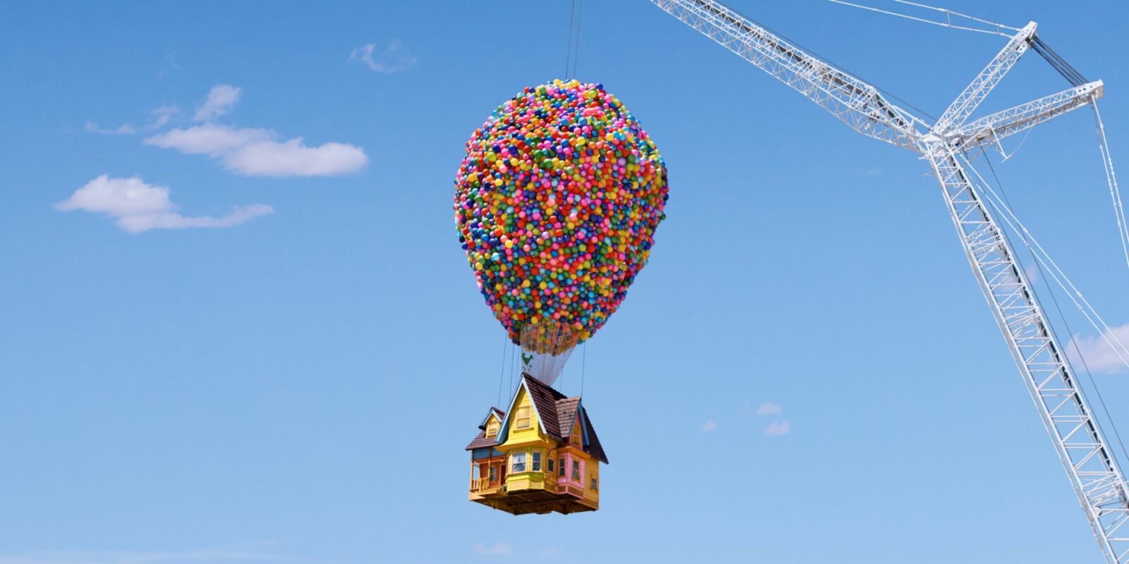 A colourful, balloon-lifted house is attached to a tall crane against a clear blue sky. The scene conveys whimsy and adventure with vibrant colours.