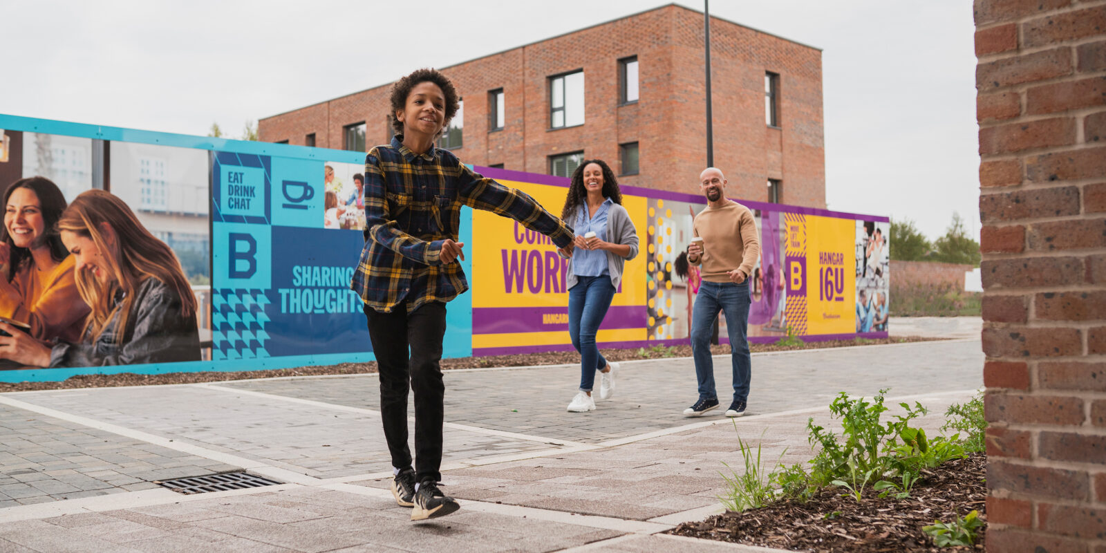 A child in a plaid shirt joyfully runs on a paved path, followed by two smiling adults. Behind them, colorful murals on a brick building create a lively urban backdrop.