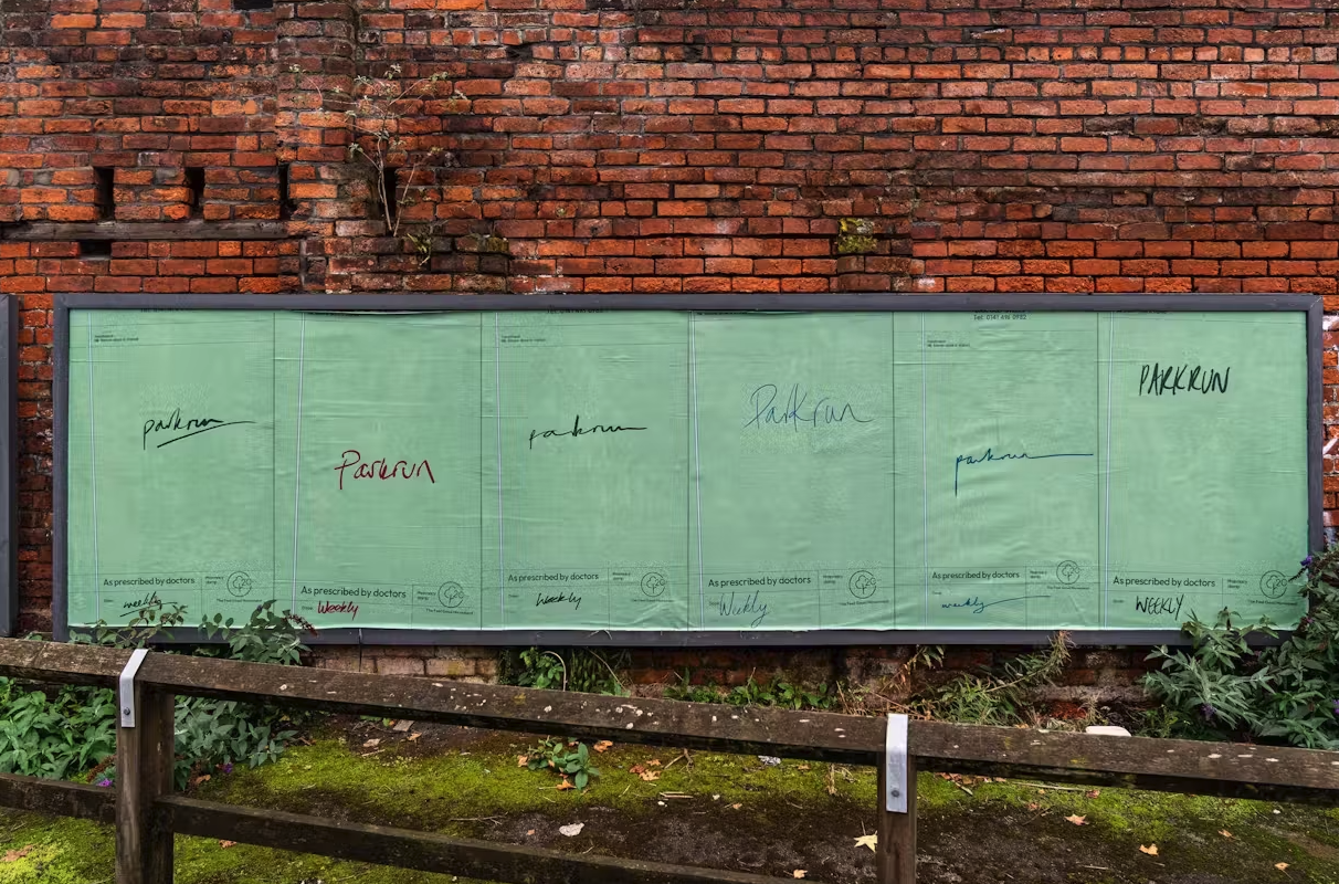 A series of green posters on a brick wall display the word "parkrun" written in various styles, each labeled "As prescribed by doctors." A rustic fence and greenery sit in the foreground.
