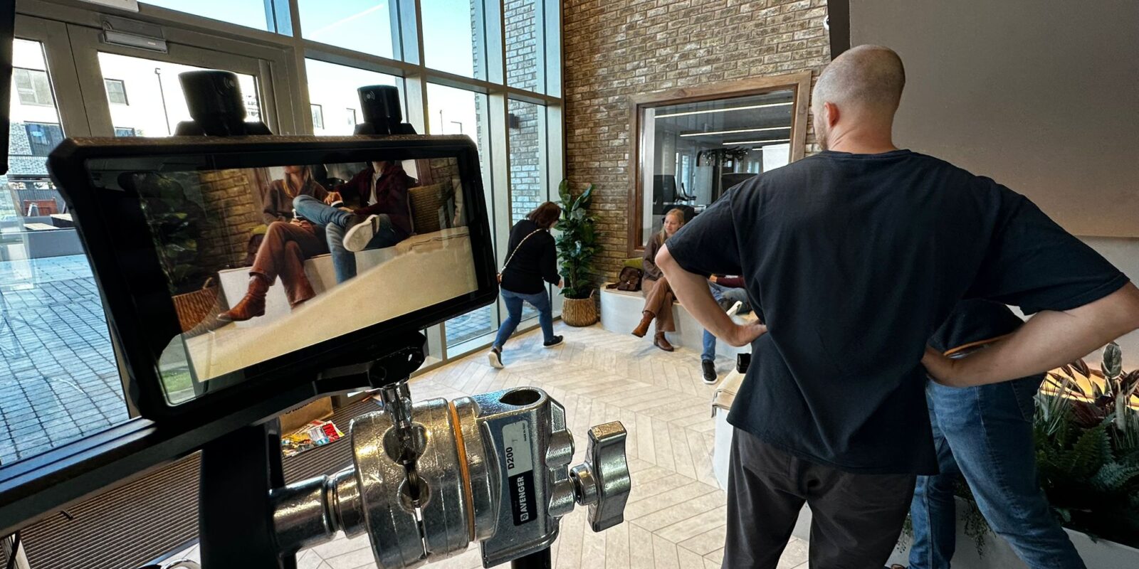 A film crew is shooting in a modern space with large windows. A camera display shows two people sitting on a couch. The atmosphere is focused and professional.
