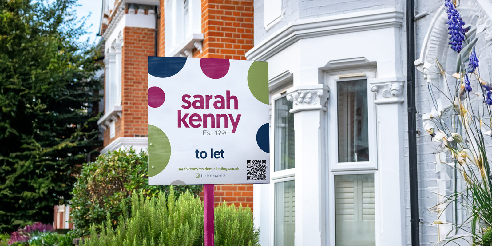 A sales board sat in front of a bay windowed house surrounded by greenery. On the sales board is the new Sarah Kenny branding. This has Sarah Kenny stacked in magenta in the top half of the board followed by to let underneath in blue smaller copy then the web address and QR code in the bottom right. Surrounding the logo are different sized playful circles in green, magenta, blue and grey