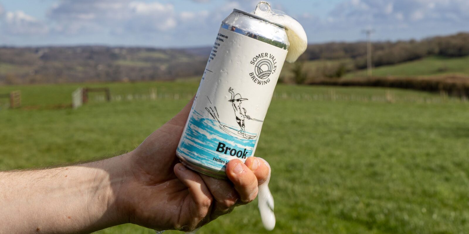 A hand holds a foaming can of beer in front of a vast green field under a partly cloudy sky. The overflowing liquid suggests a refreshing, energetic moment.
