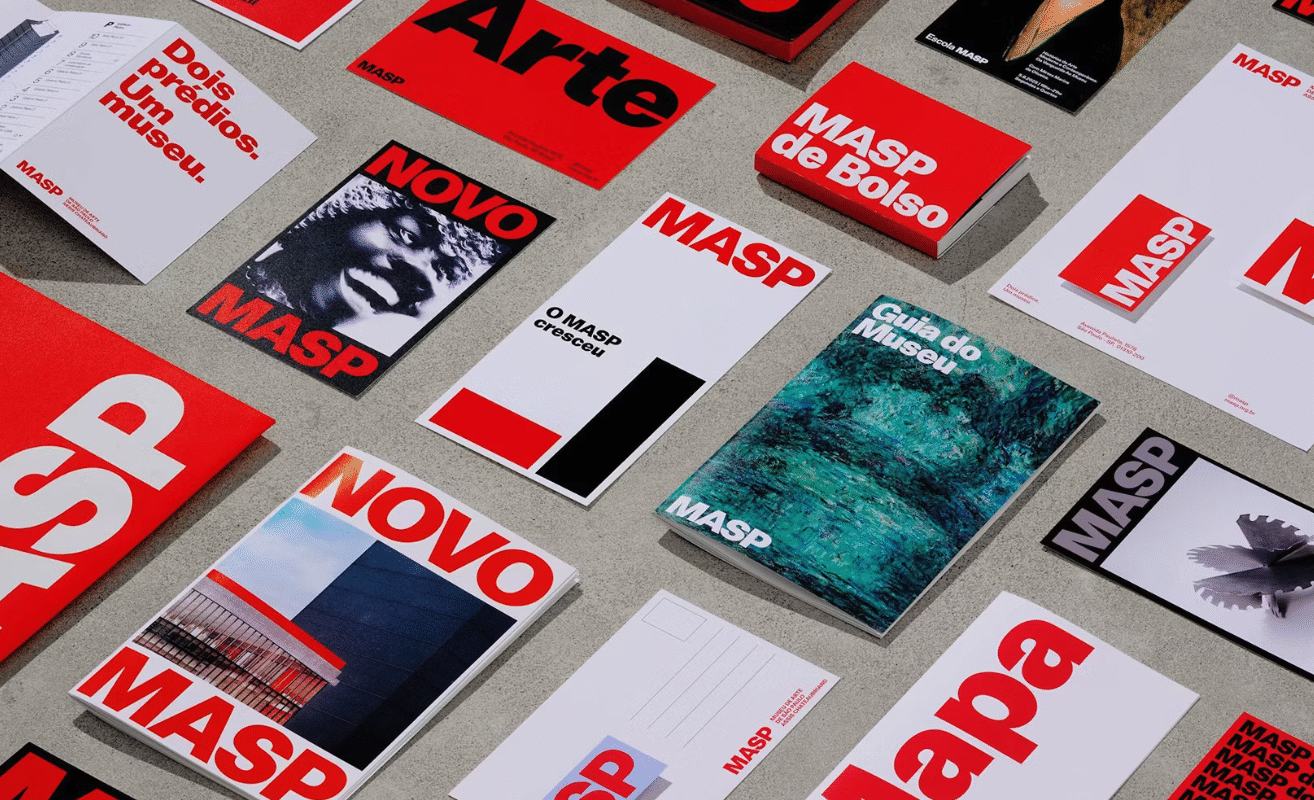 A variety of MASP-branded materials on a gray surface, featuring bold red, black, and white designs. Items include booklets, brochures, and postcards.