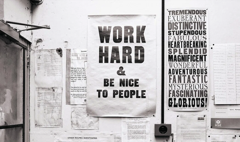 Black and white photo of a wall with motivational posters. Central text reads "WORK HARD & BE NICE TO PEOPLE," conveying inspiration and positivity.